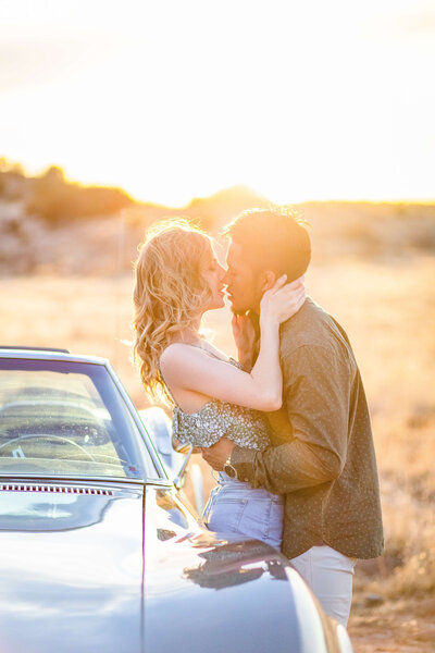 A woman and man hug as they lean up against a classic car in Sedona Arizona. The sun is setting behind them.