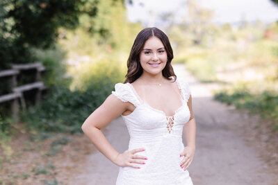 A young woman with brown hair standing on an asphalt path with greenery behind her, wearing a white, short sleeve dress with her hands on her hips, smiling at the camera in San Juan Capistrano.