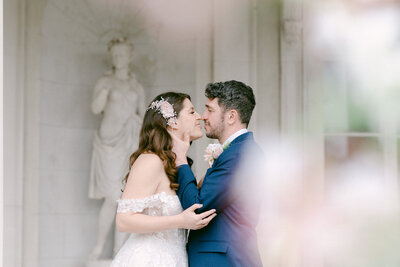 Image is of a bride and groom at Grove house, london. The groom is wearing a blue suit and has his hands placed delicately on the brides cheeks as they lean in for a kiss. The bride has a floral hair piece, a gown with off the shoulder detail sleeves and is holding the grooms elbows as they embrace.