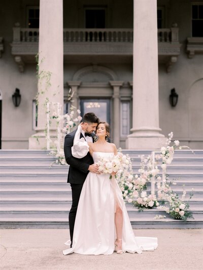 A couple stands in front of a grand building with columns, dressed in wedding attire. The groom embraces the bride from behind, who holds a bouquet of flowers—a beautiful scene orchestrated by their expert Canadian wedding planner.