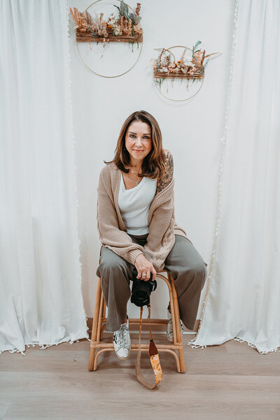 Professional photographer Nicole Mele sitting on a wooden stool in a light-filled studio, holding her camera with a warm and welcoming expression. The setting features neutral tones, boho-style wall decor, and soft natural light, reflecting her relaxed and artistic photography approach.