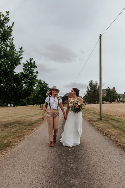 Two brides walk towards the camera down a road between two fields