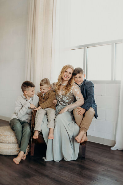 A mother cuddles with her three young sons, as they are dressed very elegantly in a neutral palette for a studio photography session in Parker Colorado. One of the sons hugs his mother's arm as the other two boys interact with each other.