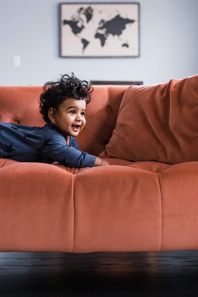 A cheerful young child with curly hair playing on a rust-colored sofa, with a world map in the background, captured by a Pittsburgh family photographer.