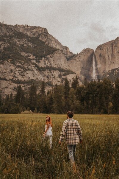 A couple walking in a field in the mountains with one in front looking back at the other.