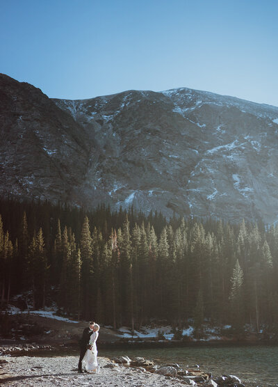 the bride and groom are drinking coffee as the sun rises. the bride is holding a navy blue canteen and the groom is holding a coffee cup with pine trees on it. they are sitting with yellow aspens and pine trees in the background. they are both smiling at each other in wedding attire.