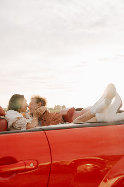 A man kissing his girlfriend while sitting in a red convertible.
