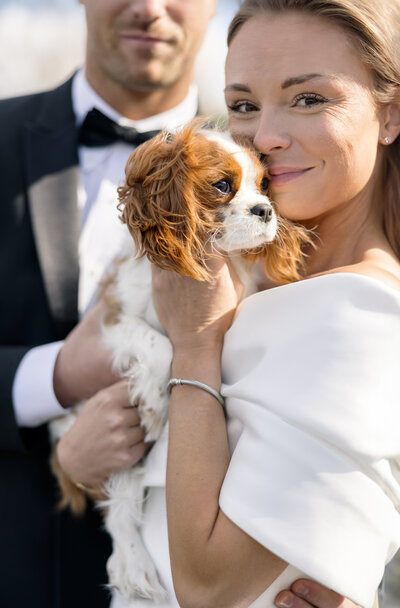 A woman in a white dress holds a small, brown and white Cavalier King Charles Spaniel, smiling at the camera. A man in a black tuxedo is partially visible beside her. The scene appears to be outdoors.