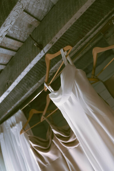 Wedding gown hangs among bridesmaid dresses on natural wood hangers