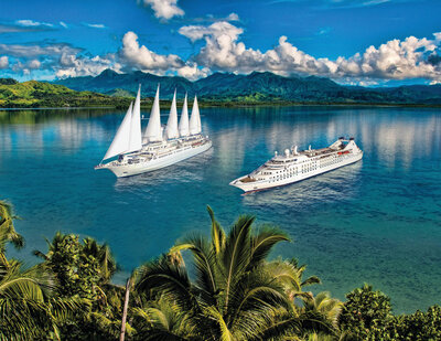 Two cruise ships, one with large white sails and the other a modern vessel, floating on clear blue water near a lush tropical coastline with mountains in the background.