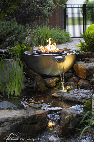 Spillway bowl with fire sitting on top of natural stones with flowing water spilling into waterfeature below