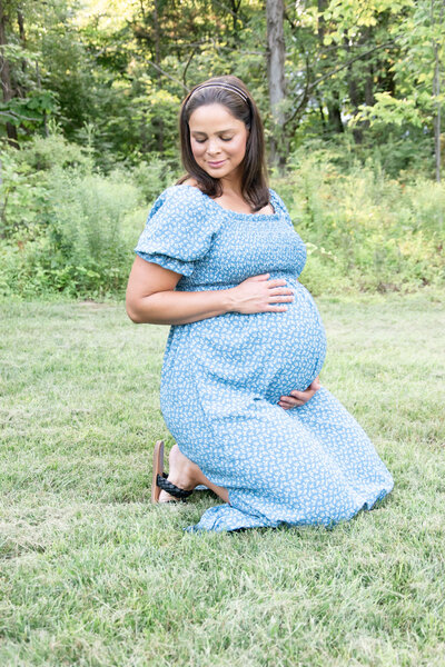Maternity mom in a blue flower dress kneeling outside.