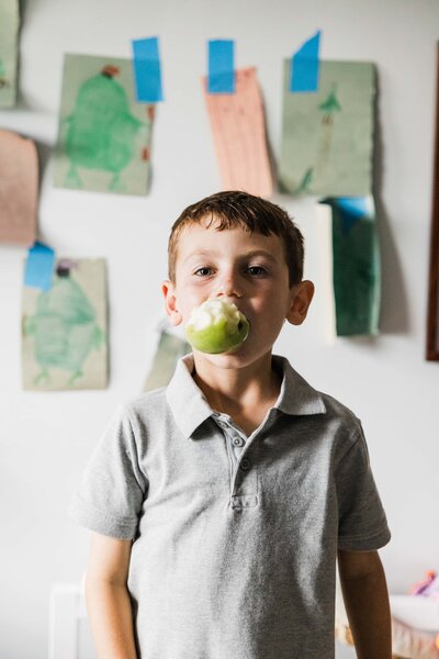 Boy with a bitten apple in his mouth standing in front of children's artwork on the wall, photographed by a renowned photographer in Pittsburgh.