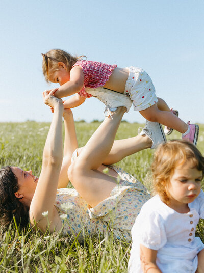 Mother lying on her back playing airplane with her daughter, captured in a loose editorial family portrait style by Emily Ambie.