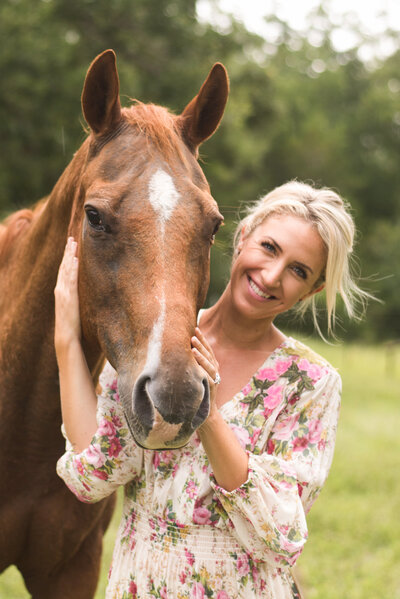 Smiling blonde girl snuggles up next to a chestnut horse looking at the camera