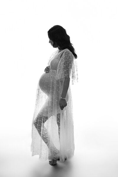 Marietta, Georgia maternity photography studio session mom in white sheer gown looking at baby bump silhouette portrait.