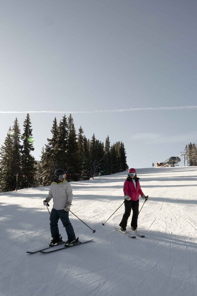 CCouple skiing together on a scenic mountain slope in Colorado, surrounded by snow-covered trees and a bright winter sky. Captured in Vail, this adventure elopement moment showcases the thrill of a winter wedding experience in the Rockies.