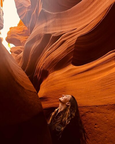 A woman standing in Antelope Canyon, surrounded by stunning sandstone walls with smooth, flowing patterns in warm red and orange hues. She gazes upward toward the light streaming in from above, capturing a sense of wonder and serenity.