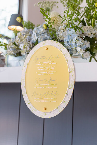 Acrylic drink menu with blue and white lettering and floral accents designed by Cove Mill Design Co. for a ‘Berry Much In Love’ wedding in Southern Maine.