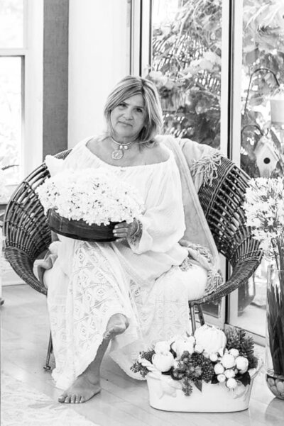 Marisol, floral and seasonal designer at Interiors by CV, seated outdoors surrounded by flowers in a white dress.