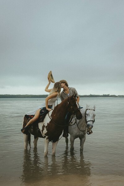 couple share a kiss on beach while riding horses