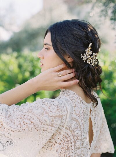 Kohnur elegant bride with natural soft makeup and intricate low updo secured with a gold crystal hair comb in a sunny garden.