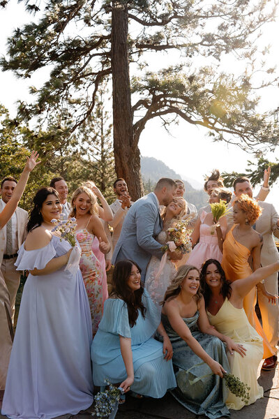 A wedding portrait of the couple with their entire wedding party. The couple is in the middle giving each other a kiss on the cheek and smiling. The wedding party is surrounding them looking excited and happy. The outfit colors are softer tones of pink, blue, orange, yellow, purple, and tan.