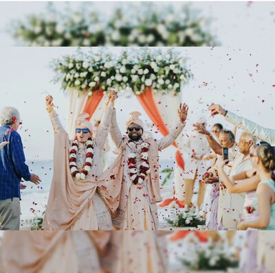 A joyful bride and groom in traditional Indian wedding attire celebrate their beach wedding, surrounded by family and friends tossing rose petals under a floral mandap.