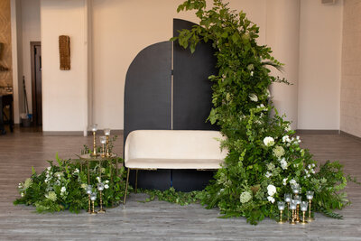 Stunning wedding photoshoot setup created by Beautifully Layered Event Rentals in Milwaukee WI. Gorgeous white accent settee small couch with a side table and black panel backdrop and greenery and white flowers cascading down from the backdrop