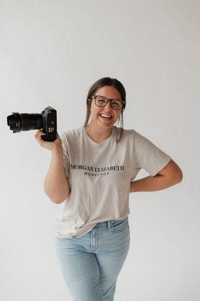 A photographer smiling with a camera in one hand an the other hand on her hip 