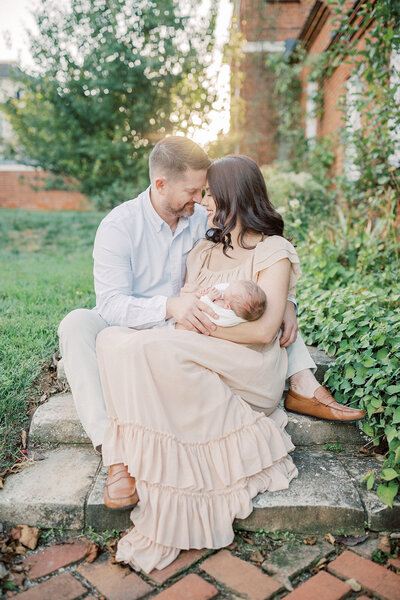 A mother and father sit on outdoor steps leaning into one another, holding their newborn baby boy during their Northern Virginia photo session in Loudoun County.