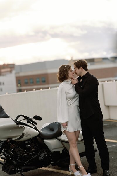 A couple stands in front of a white Harley Davidson in a pose that insinuates they are about to kiss.