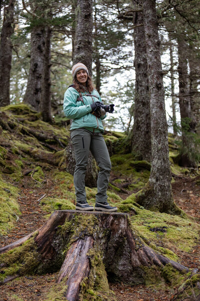 Smiling elopement photographer stands on a mossy tree stump in a dense forest, holding a camera.