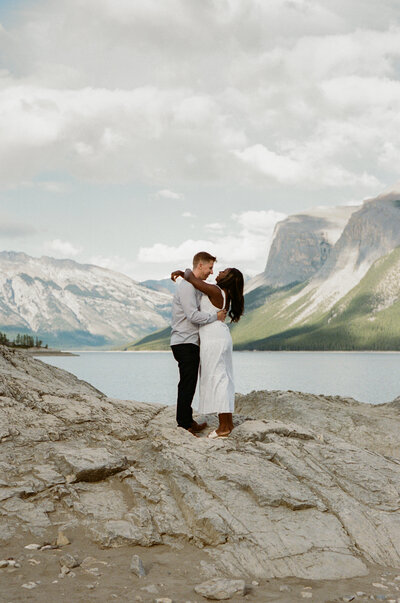 bride and groom kiss on a rocky lake shore with mountains in the background in Banff