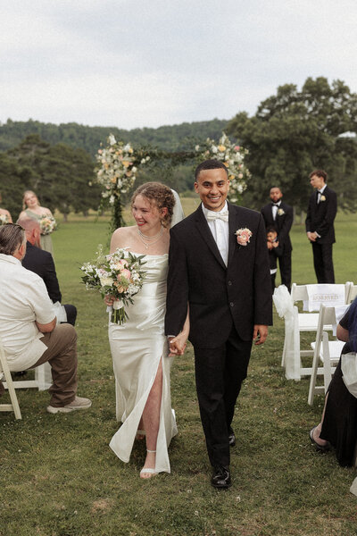 A newlywed couple walks hand-in-hand through a wedding ceremony, surrounded by guests and a beautifully decorated archway.
