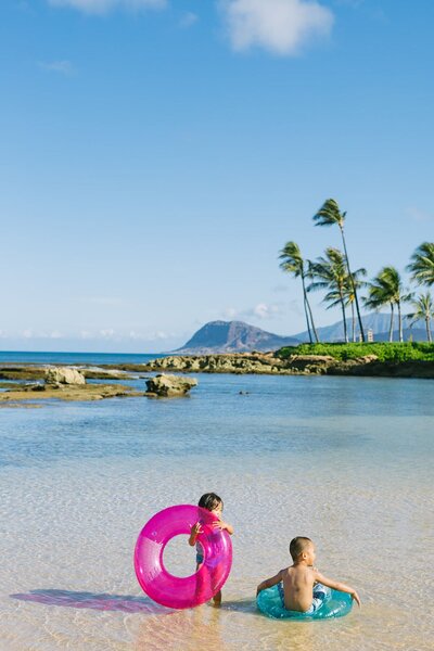 Two kids playing with brightly colored water raft sit on the beach.