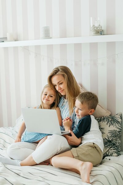 A mom sitting with her laptop on her bed with two children by her side. 