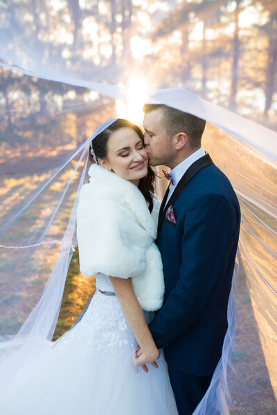 Romantic moment of bride and groom with veil over them, groom kissing her temple, eyes closed, sunset casting a warm glow behind them.
