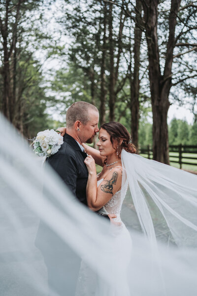 A close-up, romantic portrait of a bride and groom embracing, with the bride's veil flowing around them and a treelined backdrop at The Gentry Farm in Danville, Virginia, captured by Quinn Photography LLC.