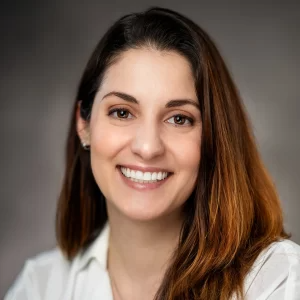 Professional headshot of woman in gray suit jacket smiling warmly against dark gray background new jersey.