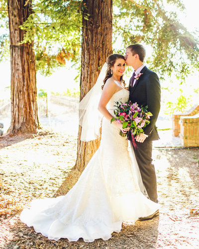 Groom kisses Bride on the forehead as she smiles at the camera holding her bouquet