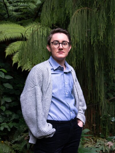 professional headshot for a young queer person, they wear a button down and cardigan with a pride pin and stand against a backdrop of greenery at the Christchurch Botanic Gardens in New Zealand.