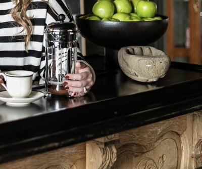 Ashleigh C. Henry, life coach and author, is wearing a black and white striped sweater, holding a vintage coffee mug, near a french press, at The Blind Tiger in North Carolina, by bright green apples at a vintage bar coffee bar.