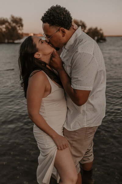 A couple share an intimate kiss by lake as the sun sets. 