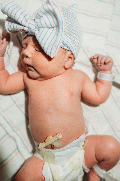A newborn baby, with a cute bow on her head, lays in the baby warmer under a light and looks very content