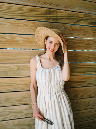 Kilee in a white dress and straw hat in front of a wood wall