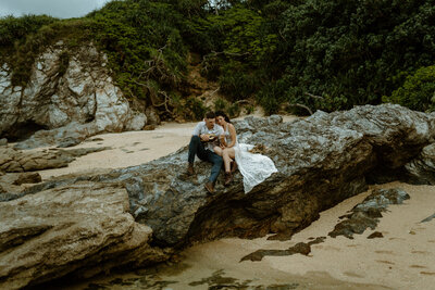 N & M sitting on a rock on the beach next to one another, playing a ukelele