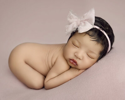 20 day old baby girl wearing a pink headband and sleeping on belly against a pink blanket covering a beanbag. 