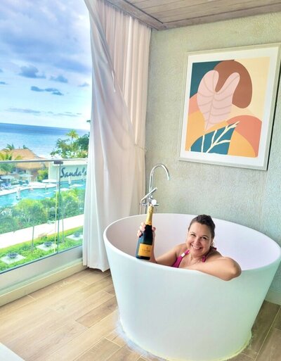Woman honeymoon planner in tub on balcony overlooking beach at Sandals and holding champagne bottle