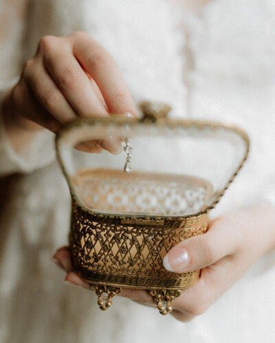 A close-up of a bride’s hands holding a gold vintage jewelry box with a delicate earring inside.
